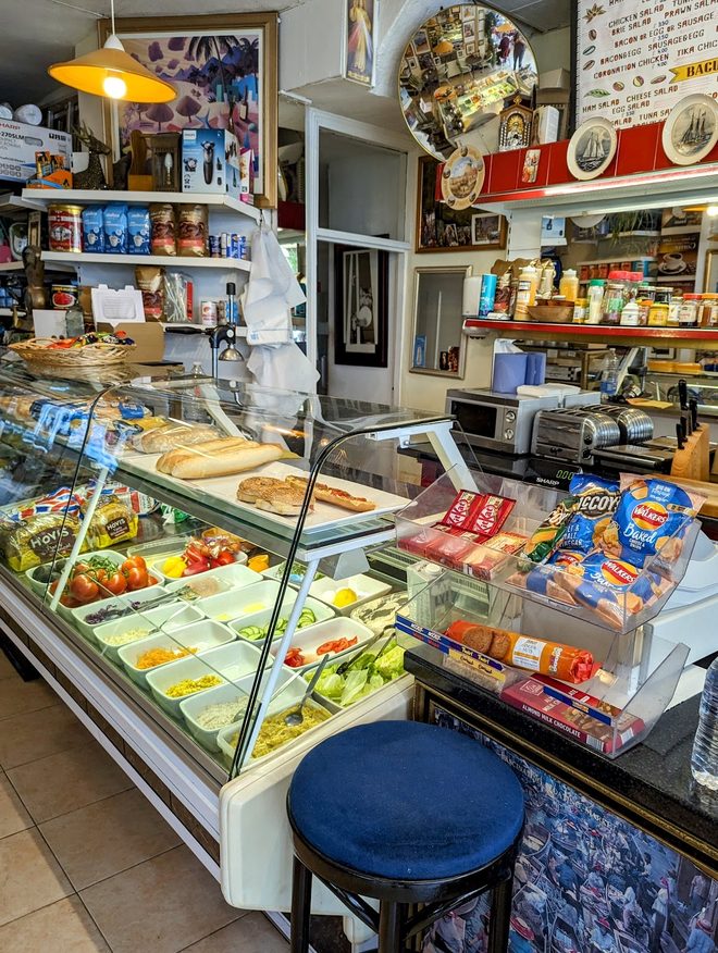 The serving counter with baguettes, fresh fruit and chilled goods, shelves of groceries behind