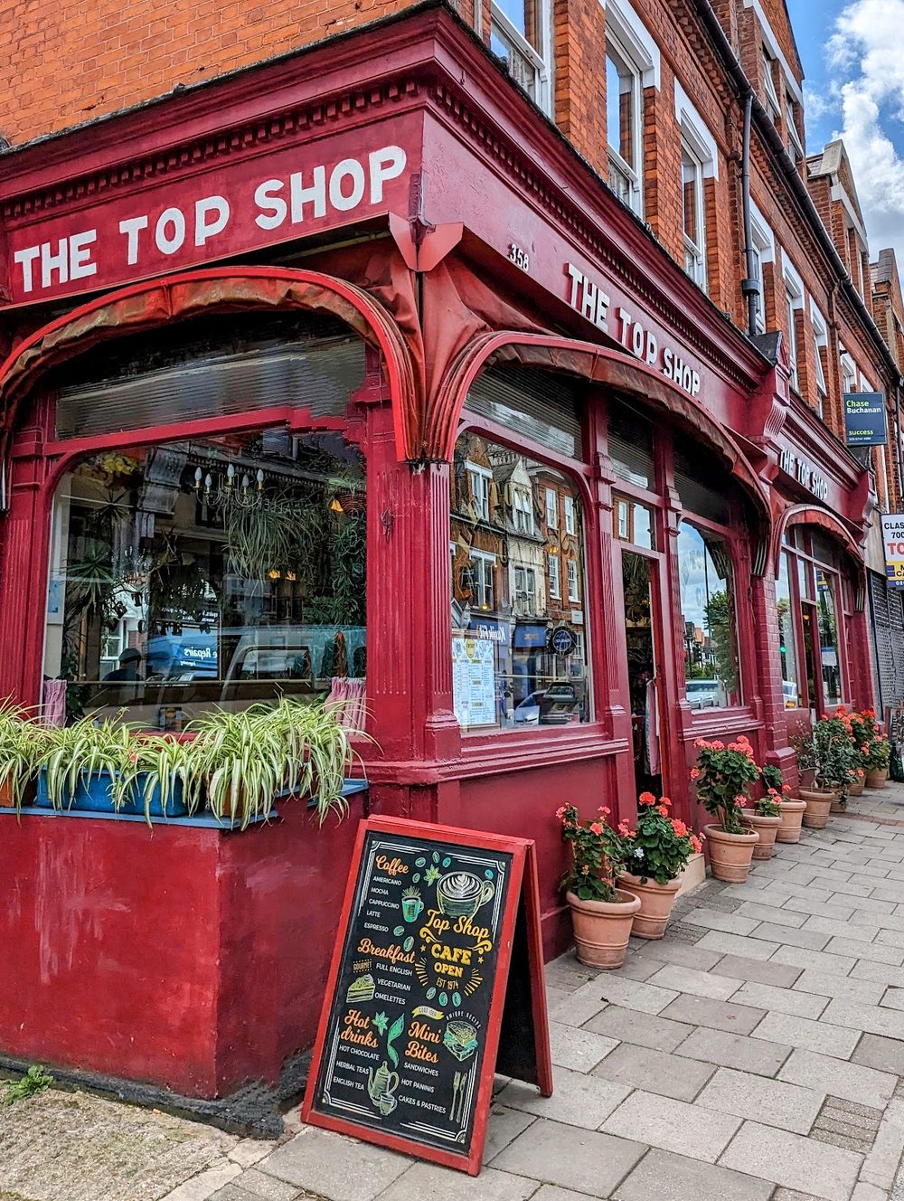 The red-painted corner shopfront seen from the pavement, with potted plants and a chalkboard outside