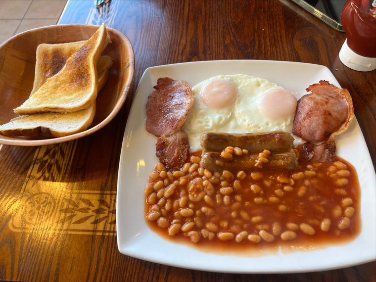 A full English breakfast on a white plate with a bowl of buttered toast